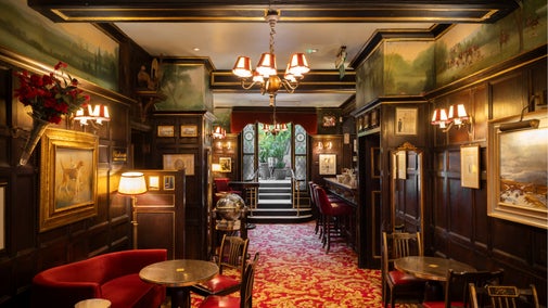 View through an elegant cocktail bar with dark wood panelling, red upholstered seating, framed artwork, and ornate lighting, leading towards a bright glass-roofed winter garden filled with greenery.